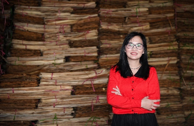 A person in red shirt and black skirt standing in front of stack of wood  Description automatically generated