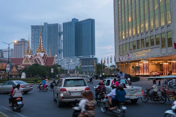 Dòng người đi qua tòa nhà Quốc hội Campuchia (bên trái) và NagaWorld (phải), sòng bạc duy nhất được cấp phép tại Phnom Penh. Phía sau là hai tòa nhà thuộc dự án căn hộ Bridge lớn và cao nhất Cam-pu-chia